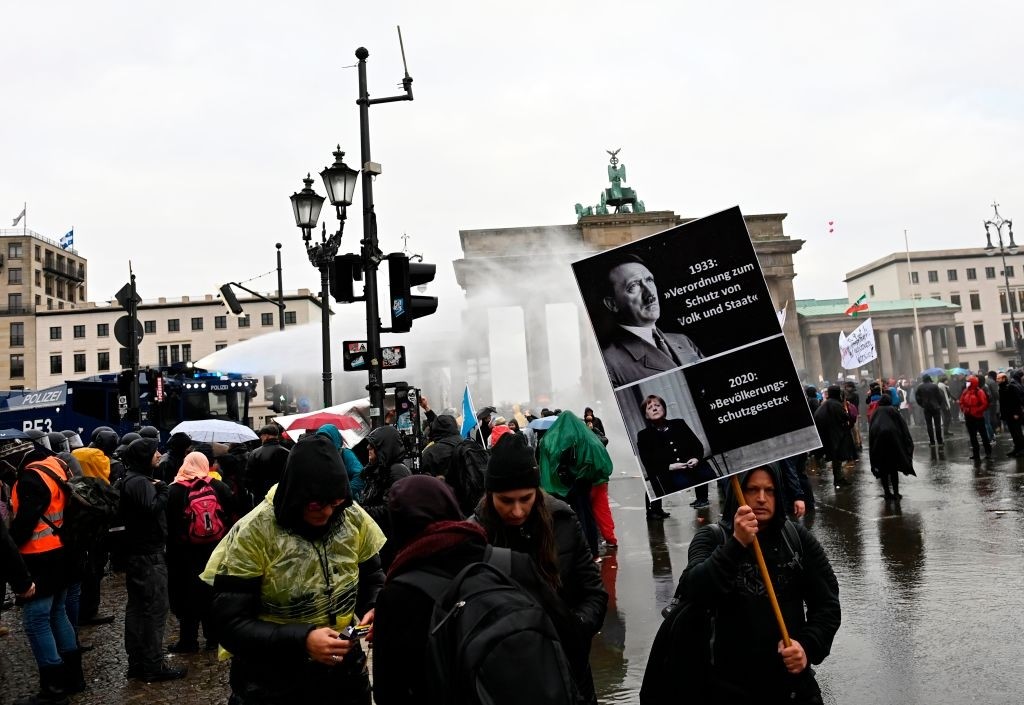 A demonstrator holds up a poster comparing Adolf Hitler's 1933 enabling act with the citizen protection law of German Chancellor Angela Merkel's government as police use a water cannon to disperse protesters demonstrating against measures imposed by the German government to limit the spread of the novel coronavirus, on November 18, 2020 at the Brandenburg Gate in Berlin. - After repeated warnings for the crowd to put on face coverings went unheeded, police said they would take action to clear the protest and "detain violators". Around 5,000 activists massed at the Brandenburg Gate, after the German government banned rallies outside parliament because of fears of violence. The demonstration mirrored similar protests seen across Europe against restrictions opponents see as a violation of their civil rights despite government warnings about the need to stop the spread of infections. (Photo by Tobias SCHWARZ / AFP) (Photo by TOBIAS SCHWARZ/AFP via Getty Images)